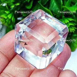 Pode incluir: Uma gema clara e facetada com um design geométrico. O cristal é segurado em uma mão, com folhagem verde ao fundo. O logotipo ParigemGifts é visível.
