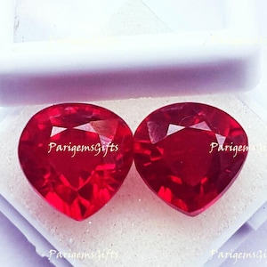 May include: Two heart-shaped, faceted, deep red gemstones. The gems are displayed on a white surface inside a white container. The text "ParigemsGifts" is visible on the gems. These gems are suitable for jewelry making.
