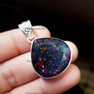 May include: A teardrop-shaped pendant with a faceted black opal gemstone. The opal displays a vibrant play-of-color with flashes of red, orange, green, and blue. The pendant is set in a silver bezel and has a decorative bail. The text "ParigemsGifts" is visible.