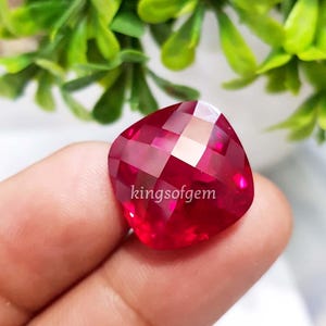 May include: A faceted, cushion-cut ruby gemstone. The gem is a deep red colour with multiple facets that reflect light. The text "kingsofgem" is visible on the gem. Green foliage is in the background.
