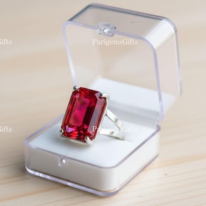 May include: A large, rectangular, ruby-red gemstone ring set in a silver band, displayed in a clear plastic box with a white interior. The ring's gemstone is the focal point, catching the light and showcasing its vibrant color.