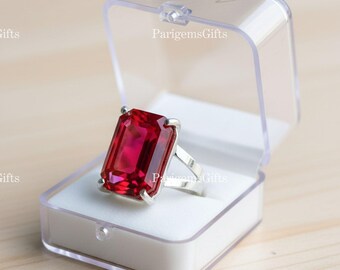 Peut inclure: Bague avec une grosse pierre rouge rubis rectangulaire, montée sur un anneau argenté, présentée dans un écrin transparent. La pierre rouge vif attire l'attention, mettant en valeur le design audacieux de la bague.