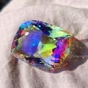 May include: A large, faceted gemstone with a vibrant display of colors, including blues, greens, and purples. The gem is cut with multiple facets, creating a sparkling effect. The gemstone is set against a light gray fabric.