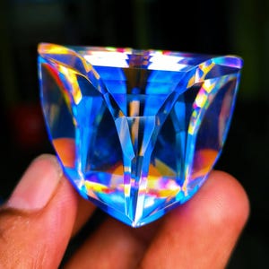 May include: A faceted, triangular-shaped glass prism featuring a vibrant blue hue with internal light refraction creating rainbow-colored highlights across its geometric, multi-angled surface, showcasing sharp edges and clear transparency.