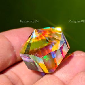 May include: A faceted, multi-colored crystal gemstone with a geometric shape. The crystal reflects a spectrum of colors, including yellow, orange, red, and blue. The gemstone is held in a hand, with a green background. The text "ParigemsGifts" is visible.