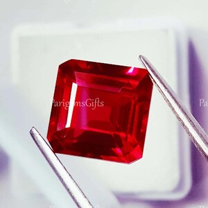 Könnte beinhalten: Ein quadratisch geschliffener Rubin, gehalten von einer silbernen Pinzette. Der Rubin hat eine tiefe, rote Farbe und eine polierte Oberfläche. Das Bild enthält den Text "ParigemsGifts".