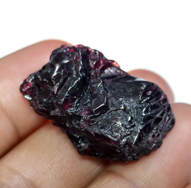 May include: A rough, uncut garnet gemstone with a deep, dark red color. The surface of the stone has a textured, crystalline appearance, with areas that reflect light. The gemstone is held in a hand.
