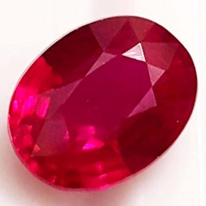 May include: A faceted, oval-cut ruby gemstone. The gem is a deep red hue, with multiple facets that catch the light. The gemstone is set against a white background, accentuating its brilliance and clarity. This ruby could be used in jewellery.