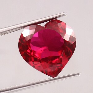 Può includere: Una gemma sfaccettata a forma di cuore di un colore rosso intenso e vibrante. La pietra è tenuta da una pinzetta in metallo color argento, che mette in risalto la sua superficie chiara e riflettente e i precisi tagli geometrici.