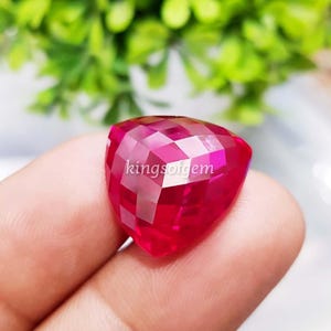 May include: A triangular, ruby-red gemstone with multiple facets. The gem is cut to reflect light, creating a sparkling effect. The text "kingsofgem" is visible on the gem. The background is blurred, with green foliage.