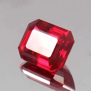 May include: A vibrant, deep red gemstone cut into a rectangular emerald shape with faceted edges. The stone exhibits high clarity and a brilliant luster, reflecting light across its polished surfaces, creating a sharp contrast against the light background.