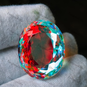 May include: A large, faceted gemstone with a vibrant display of colors, including red, teal, and green. The oval-shaped stone is held in a white glove, showcasing its brilliance. The text "Parigems Gifts" is visible on the glove.