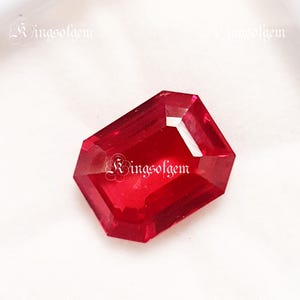 May include: A vibrant, deep red gemstone with a rectangular cut. The gem's facets catch the light, producing a bright sparkle. The gemstone is set against a white backdrop. The text "RingsofGem" is visible in the image.