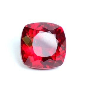 Pode incluir: Uma pedra preciosa de cor vermelha vibrante com lapidação almofada, apresentando um formato quadrado com cantos arredondados e múltiplas facetas que refletem a luz para criar uma aparência brilhante e cintilante na superfície.