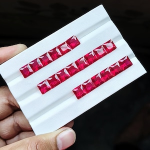 Peut inclure: Une collection de dix-sept pierres précieuses rouge vif, taillées en carré, disposées en trois rangées sur une carte de présentation blanche. Chaque pierre présente une surface à facettes qui reflète la lumière, révélant une teinte cramoisie.