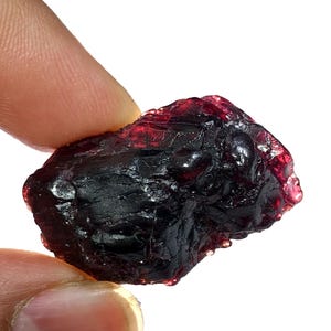 May include: A rough, dark red garnet gemstone with a textured surface. The natural stone has a deep, rich color and is held between two fingers against a white background. The gemstone is a deep, dark red.