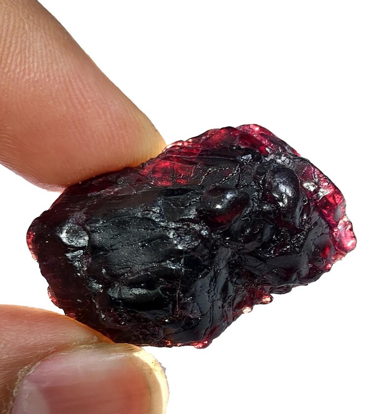 May include: A rough, dark red garnet gemstone with a textured surface. The natural stone has a deep, rich color and is held between two fingers against a white background. The gemstone is a deep, dark red.