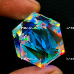May include: A multi-faceted gemstone with a brilliant display of colors, including blues, greens, and yellows. The gem is held between fingers, showcasing its intricate cut and reflective surfaces. The text "ParigemsGifts" is visible on the gem.