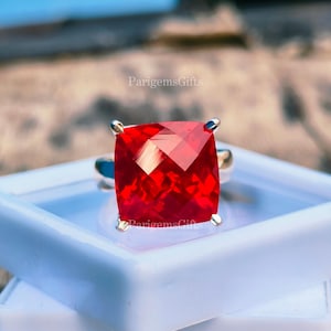 May include: A silver ring featuring a large, square-cut, vibrant red gemstone. The ring is presented in a white plastic container. The text "ParigemsGifts" is visible on the image.
