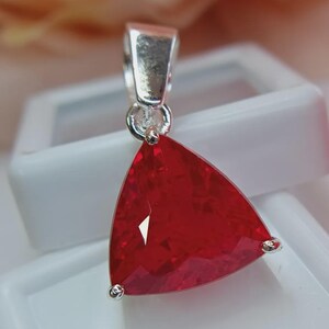 May include: A triangular pendant featuring a faceted, deep red gemstone set in a polished silver-toned metal frame. The stone has a brilliant cut with multiple reflective surfaces, and the pendant includes a silver-toned bail at the top for attachment.