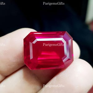 May include: A vivid, rectangular-cut ruby gemstone. The gem is a deep red colour with a polished surface. The text "ParigemsGifts" is visible on the image. The gemstone is held in a hand.