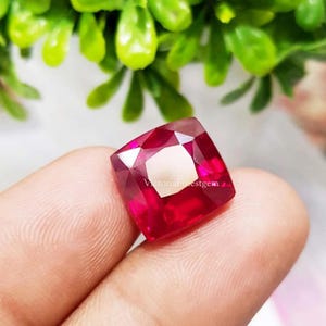 Könnte beinhalten: Ein leuchtend roter Edelstein im Kissenschliff mit einer facettierten Oberfläche, die das Licht reflektiert. Der Stein hat eine quadratische Form mit abgerundeten Ecken und zeigt einen tiefen, gesättigten Karmesinton. Der Text VictoriaFinestgem ist sichtbar.