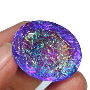 May include: A large, oval gemstone with a cut surface. The gem exhibits a vibrant array of colours, including shades of purple, blue, and green, creating an iridescent effect. The gemstone is held in a hand.