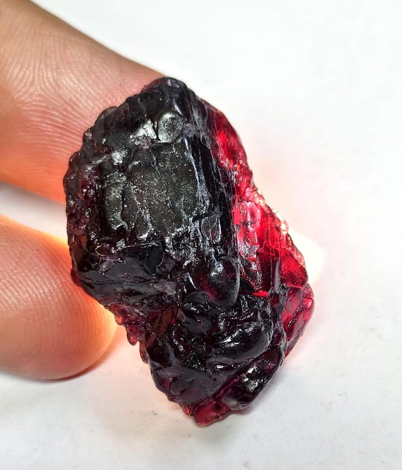 May include: A raw, uncut gemstone with a deep, dark red hue. The surface is rough and textured, with areas of near-black and translucent red. The gemstone is held between fingers, showcasing its natural, unpolished state. The overall appearance suggests a natural mineral specimen.