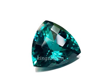 May include: A triangular, faceted gemstone with a deep teal color. The gem has a polished surface that reflects light, creating a sparkling effect. The text "kingsofgem" is visible on the gem's surface. The gem is set against a white background.