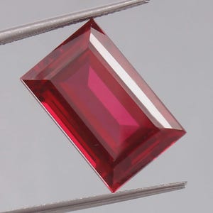 May include: A rectangular, faceted gemstone with a deep, vibrant red colour. The stone features sharp, clean edges and a polished surface that reflects light, held securely by a pair of metal tweezers against a neutral, light grey background.