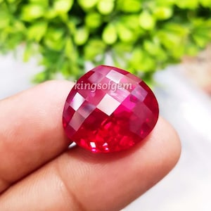 May include: A heart-shaped, ruby-red gemstone with multiple facets. The gem is held between fingers, with the text "kingsofgem" visible on the surface. The facets reflect light, creating a sparkling effect.