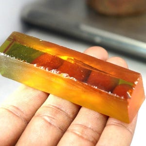 Puede incluir: Una pastilla de jabón rectangular de color ámbar con esferas rojas incrustadas. El jabón tiene una cualidad translúcida, con burbujas visibles y un ligero tono verde en los bordes. La pastilla se sostiene en una mano.