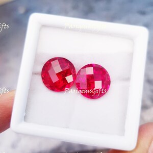 Könnte beinhalten: Zwei runde, facettierte, leuchtend rote Edelsteine werden in einer weißen quadratischen Box präsentiert. Die Edelsteine haben einen Brillantschliff, der das Licht reflektiert. Das Bild ist gut beleuchtet und hebt die Farbe und Klarheit der Steine hervor. Der Text "ParigemsGifts" ist sichtbar.