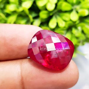 May include: A faceted, heart-shaped, ruby-red gemstone. The gem has a checkerboard pattern of facets and the text "kingsolgem" is visible on the surface. The background is blurred green foliage.