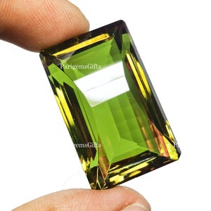 Peut inclure: Une pierre précieuse de coupe rectangulaire, d'un vert vif, rehaussée de tons dorés et bordeaux. La gemme est tenue entre les doigts, mettant en valeur ses facettes et sa clarté. Le texte "ParigemsGifts" est visible.