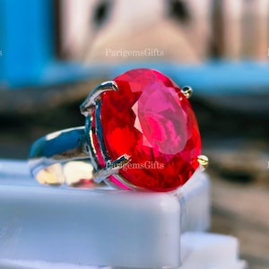 May include: A silver ring featuring a large, oval-cut, vibrant red gemstone. The gemstone is the focal point, set in a silver band with four prongs. The ring is displayed on a white surface, with the text "ParigemsGifts" visible.