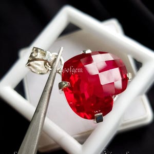 Puede incluir: Un colgante de piedra preciosa roja facetada en forma de corazón con una fianza plateada. La piedra preciosa está sujeta por cuatro garras plateadas. El colgante se muestra dentro de una caja cuadrada blanca, con unas pinzas sujetando el colgante.