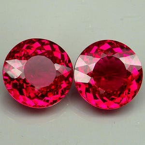 Könnte beinhalten: Zwei runde, facettierte Edelsteine in tiefroter Farbe. Die Edelsteine haben einen zentralen, helleren Bereich und reflektieren das Licht mit einem brillanten Glanz. Die Edelsteine sind vor einem neutralen Hintergrund platziert.