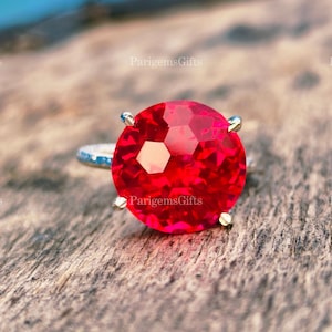 May include: A silver ring featuring a large, faceted, round, bright red gemstone. The gemstone is held in place by four silver prongs. The ring band has a textured design. The ring is set against a wooden surface.