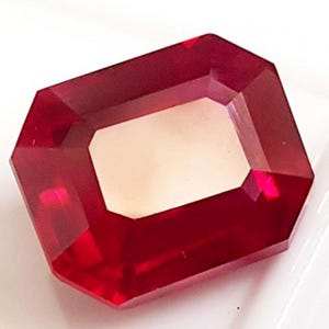 May include: A faceted, rectangular ruby gemstone with a bright red hue. The gem has a central, lighter-colored rectangular facet. The cut of the stone creates multiple reflective surfaces, enhancing its brilliance. This gemstone is suitable for jewelry making.