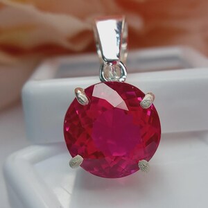 May include: A round, faceted, vibrant red gemstone pendant set in a polished silver-toned metal prong setting. The circular stone features a brilliant cut with multiple facets that reflect light, held securely by four small, textured silver prongs.