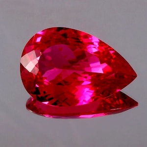Op de afbeelding: Een gefacetteerde, peervormige edelsteen met een levendige rode kleur. De edelsteen is zeer reflecterend en vertoont meerdere facetten die licht vangen en weerkaatsen. De steen ligt op een reflecterend oppervlak, wat de glans versterkt.