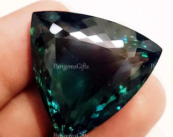 May include: A triangular, faceted gemstone with a deep teal and dark blue color gradient. The gem has a polished surface that reflects light, creating bright highlights. The text "ParigemsGifts" is visible on the gem's surface.
