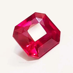 Könnte beinhalten: Ein facettierter, achteckiger Rubin-Edelstein mit einem quadratischen Ausschnitt in der Mitte. Der Edelstein hat eine tiefe, leuchtende rote Farbe, wobei mehrere Facetten das Licht einfangen. Der Edelstein ist vor einem weißen Hintergrund platziert.