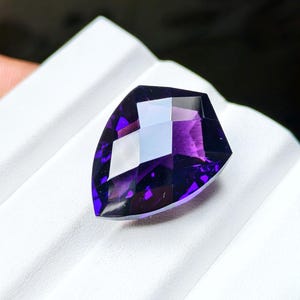 May include: A deep purple, shield-shaped gemstone with a faceted surface. The stone features a pointed bottom and a flat top edge, displaying a rich violet hue with light reflections across its polished, geometric, multi-faceted cut surface.