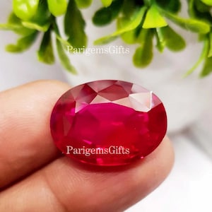 Könnte beinhalten: Ein facettierter, oval geformter Rubin-Edelstein. Der Edelstein hat einen leuchtenden Rotton, wobei mehrere Facetten das Licht einfangen. Der Text "ParigemsGifts" ist auf dem Edelstein aufgedruckt. Grünes Laub ist im Hintergrund zu sehen.