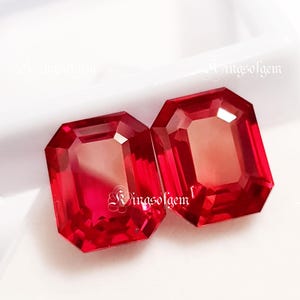 Puede incluir: Dos gemas rojas facetadas de corte rectangular. Las gemas tienen múltiples facetas que reflejan la luz, creando un efecto brillante. Las gemas están sobre un fondo blanco. El texto "WingsofGem" es visible en el fondo.