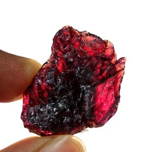 May include: A rough-cut, deep red gemstone with a translucent quality. The stone has a dark, almost black, interior and a vibrant, ruby-like hue in the lighter areas. The edges are uneven, showcasing its natural, raw form.