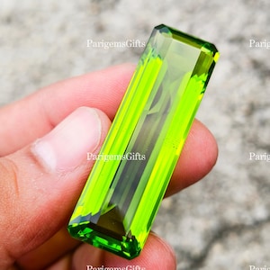 Può includere: Una gemma rettangolare, sfaccettata, verde brillante. La gemma è tenuta in mano, mostrando la sua chiarezza e il suo colore vibrante. Le sfaccettature della gemma catturano la luce, creando un effetto scintillante. La gemma è un pezzo vivido e accattivante.