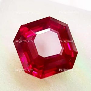 May include: A faceted, octagonal ruby gemstone with a bright red hue and a central, lighter-colored facet. The gem is set against a soft, white background. The text "ParigemsGifts" is visible in the corners.